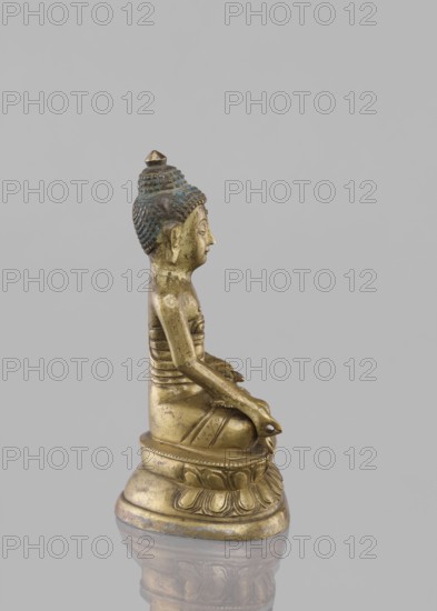 Buddha sitting on a lotus throne, figurine, Chinese culture, Chinese art, Buddha iconography, Buddhist art, lotus throne iconography, Harbin, China, Qing 1644–1911, ushnisha, 19th century, Tibet, sculpture, height 14.4 cm, A small metallic statue of a seated figure viewed in profile against a plain light gray background, showing a golden-brass tone overall with areas of darker patina and greenish-blue oxidation on the hair and upper head; the figure sits cross-legged on a rounded decorative base with petal-like carvings, hands resting in the lap, elongated earlobes, a detailed textured hairstyle topped by a small finial, and smooth sculpted contours on the torso and limbs, with reflections of the base visible on the glossy surface beneath; visible colors include gold, brass, greenish-blue, dark brown, and light gray.