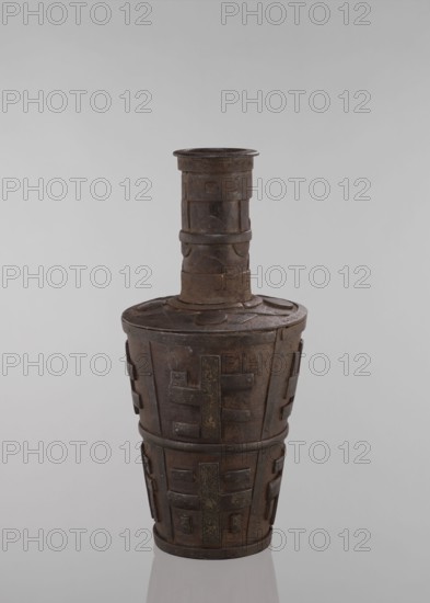 Jug, vessels, Tibetan culture, vases, meander ornament, symbols, Buddhist art, vessel tapering towards the bottom, 18th–19th century, Tibet place of origin ?, vessel, height 46.3 cm, An upright vessel with a tall narrow neck and a wider rounded body, made of a dark brown, rust-toned metal with areas of lighter brown and black patina, featuring raised rectangular and circular decorative panels and bands encircling the neck and body; the surface shows textured and worn areas, and the object stands on a reflective pale gray surface against a smooth light gray background.