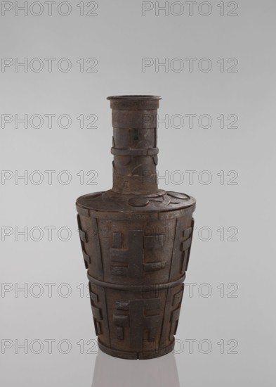 Jug, vessel tapering towards the bottom, Tibetan, 18th–19th century, ceramic vessel, height 46.3 cm; features meander ornament, symbolic motifs, Buddhist art, vases., A tall, narrow vessel with a long cylindrical neck and a wider, tapered body sits against a plain light gray background. The surface of the object appears textured and metallic with visible wear and a patina of dark brown and rust tones, along with areas of darker gray. The neck has a few raised bands and rectangular applied elements, while the shoulder and body display repeated raised geometric motifs and rectangular panels in the same dark brown and gray palette. The top rim is slightly flared and shows the same worn metallic coloration, and the base sits on a reflective surface that picks up faint gray reflections.