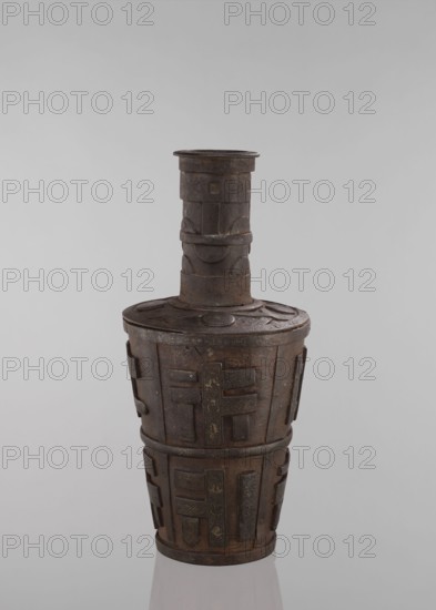 Jug, vessels, Tibetan, vases, meander ornament, symbols, Buddhist art. Vessel tapering towards the bottom. 18th–19th century, Tibet, vessel. Height 46.3 cm., The image shows a tall, narrow vessel with a long cylindrical neck and a wider, rounded body, rendered in dark brown metal with areas of lighter brown and subtle greenish patina; the surface features raised geometric and rectangular relief panels and bands that wrap around the body and neck, and a few circular disc motifs on the lid area; the vessel rests on a reflective pale surface against a smooth light gray background.