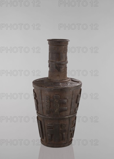 Jug, vessels, Tibetan, vases, meander ornament, symbols, Buddhist art, vessel tapering towards the bottom, 18th–19th century, Tibet, vessel, height 46.3 cm, A tall, cylindrical vessel with a narrow neck and wider body, made of a dark brown metal with a weathered, slightly rusty surface; the neck has a few raised bands and carved geometric patterns, and the body is decorated with several raised rectangular and angular relief pieces that create a repeating motif around the form; the top rim is flat and the shoulder is slightly rounded with circular raised discs, while the base tapers inward and rests on a reflective surface; visible colors include dark brown, rusty brown, and hints of black against a pale gray background.
