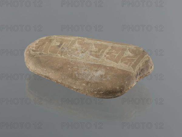 Mani stone, Tibetan Buddhist art, Tibetan culture, Buddhism, mantras, inscriptions, ritual stone, 19th/20th century, Tibet, cult object; weight 1.04 kg, height 16.7 cm, width 10.5 cm, A smooth, rounded flat stone rests on a reflective gray surface, showing a worn top with lighter scratches and incised patterns. The stone is mostly tan and brown with areas of pale beige and faint white scuffing. Its edges are slightly darker brown and one end shows a small rounded notch with darker brown discoloration. The surface reflects softly below, producing a muted mirrored shape in shades of gray and brown. The background is a uniform mid-gray.