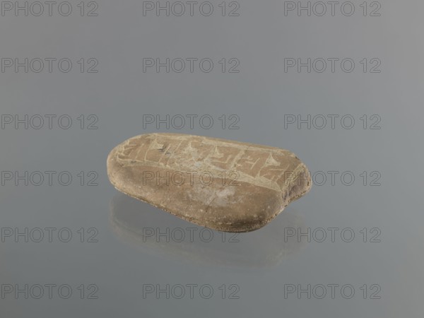 Mani stone, Tibetan Buddhist art; Tibetan culture; Buddhism; mantras; inscriptions; ritual stone; 19th/20th century; Tibet; cult object; weight 1.04 kg; height 16.7 cm; width 10.5 cm., A rounded, flat stone with a smooth surface rests on a reflective grey background; the stone is primarily light brown with areas of beige and pale tan, and it shows a lighter band of worn markings across its top surface with faint patches of darker brown and small speckles of white.