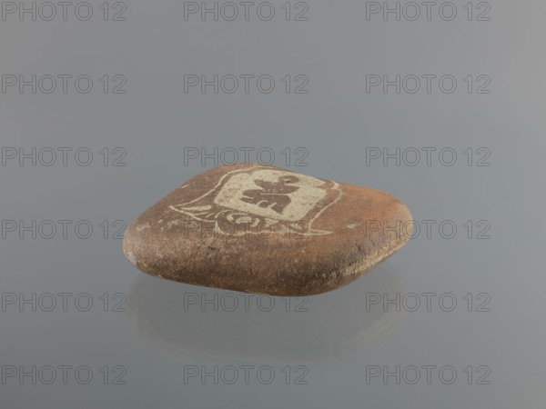 Mani stone, gift; Tibetan Buddhist art; Tibetan culture; Buddhism; mantras; inscriptions; ritual stone; 19th/20th century; Tibet; cult object; weight 1.64 kg; height 14.0 cm; width 15.5 cm, A smooth, flat brown stone with lighter beige markings on its top surface rests on a reflective gray surface, showing a soft reflection beneath; the background is a uniform gray.