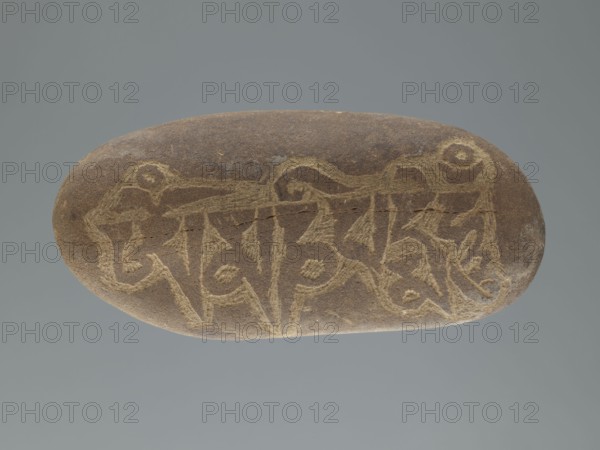 Mani stone, gift, Tibetan Buddhist art, Tibetan culture, Buddhism, mantras, inscriptions, ritual stone, 19th/20th century, Tibet, cult object, weight 0.67 kg, height 15.5 cm, width 7.5 cm, A smooth oval stone with a brown surface rests against a muted gray background; a series of shallow, lighter-colored incised lines form an intricate linear pattern across the top of the stone, creating angular and curved shapes that include spirals and pointed elements; the stone shows subtle variations in brown tones and a few paler abrasions where the incised design appears lighter.
