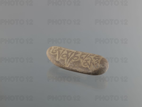 Mani stone, gift, Tibetan Buddhist art, Tibetan culture, Buddhism, mantras, inscriptions, ritual stone, 19th/20th century, Tibet, cult object; weight 0.67 kg, height 15.5 cm, width 7.5 cm, A smooth oval stone with a brown and tan surface rests on a reflective gray surface, showing a mirrored reflection beneath; the stone has lighter tan carved or etched patterns on its top and rounded ends, with the surrounding background and reflection in shades of gray and pale blue.
