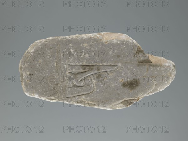 Mani stone, gift; Tibetan Buddhist mani stone with inscriptions of mantras; ritual cult object, stone slab; Tibetan, 19th–20th century; material: stone; weight 0.7 kg; height 18.3 cm; width 9.0 cm., A single irregularly shaped stone rests against a plain gray background; the stone has a muted palette of light gray with areas of beige and pale brown, mottled with small lighter flecks and a few darker gray spots. The stone’s surface is uneven with smooth worn areas, shallow grooves, and a few incised linear marks forming angular shapes. Edges of the stone are rounded in places and slightly jagged in others, showing subtle texture variations and faint patches of discoloration in beige and brown tones. The overall appearance is of a weathered, pale gray rock with beige highlights and scattered darker gray blemishes.