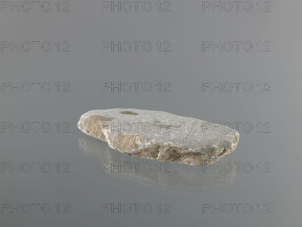 Mani stone, gift; Tibetan Buddhist ritual stone inscribed with mantras; 19th/20th century, Tibet; cult object; stone slab; weight 0.7 kg; height 18.3 cm; width 9.0 cm; Tibetan Buddhist art; Tibetan culture; Buddhism; inscriptions., A flat, irregularly shaped stone with a rough, pitted surface rests on a smooth reflective surface, showing a faint mirror image beneath it; the stone displays shades of gray, beige, light brown, and off-white, with some darker gray spots and weathered edges, and the background and surface are various tones of muted gray.