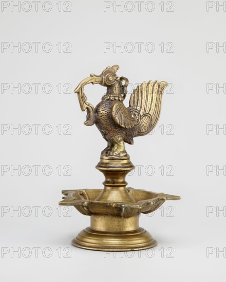 Oil lamp; round base supporting a pentagonal oil vessel with a bird figurine above. 19th century, South India Tamilnadu. Materials: oil lamp metal; iconography: birds rooster, goose, swan, fire; associated with Hindu religion, temples, rituals. Height 19.0 cm; width 13.0 cm., A metal object with a golden-brass color is shown against a plain light background; it features a sculpted bird figure with textured feathers, a raised tail with lined detailing, and a curved beak holding a decorative element, mounted on a turned pedestal that sits in a shallow scalloped dish atop a rounded base, with visible surface patina and areas of darker and lighter golden tones.