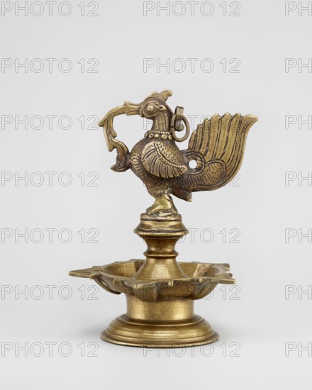 Oil lamp, round base with a pentagonal oil vessel and a bird figurine on top; 19th century South India Tamil Nadu; materials: oil lamp; subject/iconography: birds roosters, geese, swans, fire, religion, temples, Hinduism, rituals; height 19.0 cm, width 13.0 cm., A brass object shaped like a stylized bird stands on a rounded pedestal base, featuring a flared, scalloped dish at the top of the base and a turned column supporting the bird; the bird has a detailed textured body with carved feather patterns, a raised decorative collar, a curved beak with an open loop near it, and an ornate fan-shaped tail with engraved lines; the surface shows areas of brighter and darker golden tones and subtle greenish patina; the background is a plain light gray.