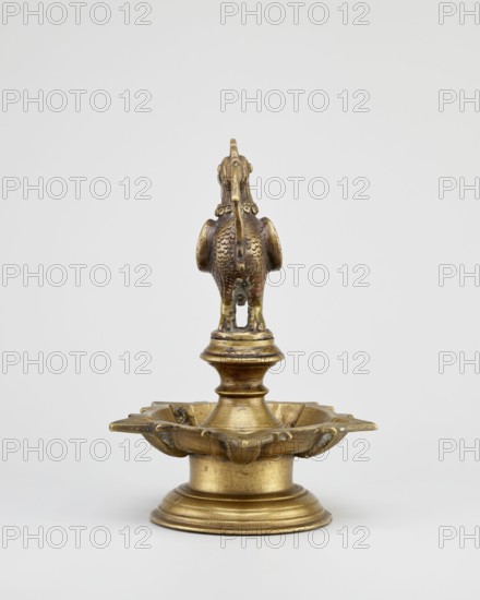 Oil lamp, 19th century, South India, Tamilnadu. Round base with a pentagonal oil vessel above which is a bird figurine; iconography includes birds roosters, geese, swans. Materials: oil lamp; function: ritual/temple use in Hinduism; height 19.0 cm, width 13.0 cm., A small metallic object sits centered on a plain light-gray background, appearing as a decorative stand with a rounded tiered base and a scalloped dish above it, finished in aged gold with darker patina in recesses; rising from the dish is a short fluted column topped by a sculpted bird figure seen from the rear, showing textured feather details, a long curved tail, and a rounded head with small protrusions, all sharing the same gold color with areas of brownish wear.