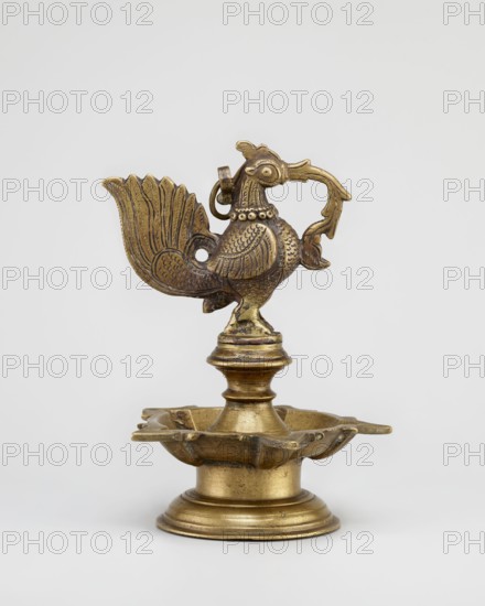 Oil lamp, gift, Indian art, Indian culture, bird iconography, fire, religion, temples, Hinduism, rituals, roosters, geese, birds, swans. Round base with a pentagonal vessel for oil; above a bird figurine. 19th century, South India, Tamilnadu. Oil lamp; height 19.0 cm, width 13.0 cm., A brass object shaped like a stylized bird perched on a pedestal above a shallow scalloped dish, the bird featuring a fan-shaped tail, patterned wings, a curved beak, and a ring near its head; the pedestal has layered round forms and the dish has petal-like edges; the colors visible are gold and darker brownish-gold tones against a light gray background.