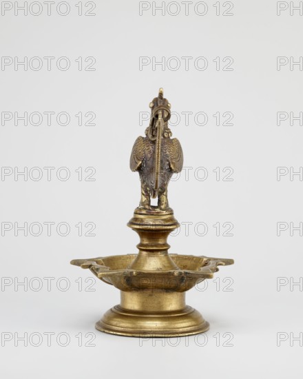 Oil lamp, Round base with a pentagonal vessel for oil topped by a bird figurine rooster/swans/geese motifs; 19th century, South India, Tamilnadu; oil lamp; Indian; bird iconography; Hinduism, temples, rituals; height 19.0 cm, width 13.0 cm., A metallic object with a worn gold and brown patina stands on a round tiered base and rises to a shallow dish with scalloped edges, topped by a central turned column supporting a sculpted bird figure with detailed feather texture and a hooked beak; the colors visible are gold, brown, and hints of darker bronze tones against a light neutral background.