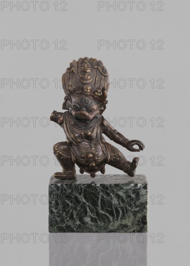 Lamaist deity with a bull's head — Yama; maker, Tibetan Buddhist sculpture/figurine; 19th century, Tibet; Tibetan Buddhist art; materials: sculpture figurine; subject: Lamaist deity with a bull's head Yama; motifs: bulls; height 12.8 cm., A small bronze-colored figurine with a dark brown patina stands in a dynamic squatting pose on a green and black marbled rectangular base; the figure has an ornate headdress with rounded protrusions, a fierce facial expression with pronounced eyes and open mouth, detailed jewelry and draped garments with decorative circular elements, and one arm bent while the other extends outward, all shown against a plain light gray background with subtle reflection beneath the base.