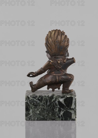Lamaist deity with a bull's head — Yama; creator, Tibetan Buddhist art; 19th century, Tibet; sculpture, figurine; materials unspecified; height 12.8 cm; subject: Lamaist deity with a bull's head Yama; function: religious/ritual figurine; inscriptions: none specified., A small bronze figure with a dark brown and golden patina shown from the back, kneeling on a polished green and black marble block; the figure has a stylized feathered or flamelike headdress with textured ridges, a short skirt with carved folds and decorative details, arm and leg bands, and one arm extended behind while the other is bent forward, all set against a plain light gray background.