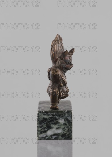 Lamaist deity with a bull's head — Yama; creator, Tibetan Buddhist art; sculpture, figurine; material not stated; 19th century, Tibet; height 12.8 cm; subject: Lamaist deity Yama depicted with a bull's head; themes: Buddhist art, Tibetan culture, bulls., A small bronze figurine with a dark brown and golden patina stands on a square green and white marbled base, showing a stylized humanoid figure with an elongated ornate headdress, a pronounced snout or mask-like face, layered clothing or armor with rounded decorative elements, and a slightly forward-leaning posture.