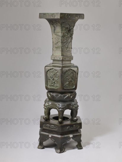 Vase-shaped stove, Chinese art, Chinese culture, unknown provenance; vase-shaped stove decorated with plant ornament, pines, phoenixes, tigers, lotus throne, gourds, dragons, sages, vipers, warriors, lions; Qing dynasty 1644–1911, 19th century, Chinese Buddhism and Taoism iconography; height 138 cm, width 42 cm, weight 65.5 kg., The image shows a tall metal object in shades of dark green and bronze with a vertical, multi-tiered structure; the top section flares outward with carved reliefs and a flat rectangular rim, the middle portion is a squared column with rounded edges featuring framed relief panels on its sides, and below that is a rounded, lotus-patterned segment supported by a pedestal of four short, curved legs with stylized animal faces; the base consists of a hexagonal platform with carved motifs standing on a decorative stand with curved cutouts and feet, all set against a plain, light gray background.
