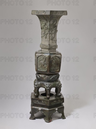 Vase-shaped stove, Chinese art, Chinese culture, unknown provenance; vases; plant ornament; Qing dynasty 1644–1911; themes: Chinese Buddhism, Taoism, Immortals; motifs: pines, phoenixes, tigers, lotus throne, gourds, dragons, sages, vipers, warriors, lions; 19th century, China; function: stove; height 138 cm, width 42 cm, weight 65.5 kg., A tall metallic object with a dark greenish-brown patina standing against a plain light gray background; the object is composed of stacked sections including a flared top with carved circular motifs, a tapered column with raised floral and organic reliefs, a squared midsection with recessed panels each containing figural reliefs, a rounded base section with lotus petal motifs, a ring supported by four short legs carved with stylized animal faces, and a hexagonal lower platform resting on four curved feet with pierced and scalloped details; surfaces show variations of green, brown, and dark bronze tones and areas of subtle sheen.