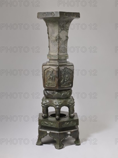 Vase-shaped stove, Chinese art, Chinese culture, unknown provenance; vase-shaped stove decorated with plants, pines, phoenixes, tigers, lotus throne, gourds, dragons, sages, vipers, warriors, lions; associated with Chinese Buddhism, Taoism, Immortals iconography; Qing dynasty 1644–1911, 19th century, China; height 138 cm, width 42 cm, weight 65.5 kg., A tall metal sculpture with a greenish-brown patina stands against a plain light gray background; it has a flared upper section with decorative raised motifs, a square midsection with carved relief panels showing figures and natural elements, a rounded base section with a carved lotus-like band and a group of bulbous supports topped by stylized animal faces, and a lower square platform supported by ornate cut-out feet, all showing varying shades of green, brown, and dark bronze.