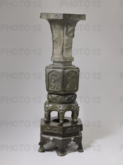 Vase-shaped stove, Chinese art, Chinese culture, unknown provenance; materials: vases; motifs: plant ornament, pines, phoenixes, tigers, lotus throne, gourds, dragons, sages, vipers, warriors, lions; iconography: Chinese Buddhism, Taoism, Immortals; period: Qing 1644–1911, 19th century; function: stove; dimensions: height 138 cm, width 42 cm, weight 65.5 kg., A tall metal vessel with a greenish patina stands on a matching stand; the vessel has a flared top section with a squared rim and a vertical neck decorated with raised ornamental designs, a squared midsection with rounded corners showing embossed figurative panels, a rounded lotus-like collar, and a four-legged base with sculpted animal-head details that sits on an ornate low platform base with curved legs. The visible colors include green, dark bronze, and hints of brown.