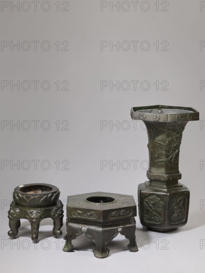 Vase-shaped stove, Chinese, Qing dynasty 1644–1911, 19th century. Porcelain/ceramic stove shaped like a vase, height 138 cm, width 42 cm, weight 65.5 kg. Decoration includes plants, pines, phoenixes, tigers, lotus throne, gourds, dragons, sages, vipers, warriors, lions; iconography related to Chinese Buddhism, Taoism, and Immortals. Function: stove. Place of origin: China., The image shows a group of three metal objects arranged on a neutral background, all with a dark greenish-brown patina. On the left is a low, round vessel with a wide rim sitting on a short tripod base with bulbous, animal-like legs and carved decorative motifs, the rim and upper surface appearing slightly darker and more worn. In the center is a hexagonal stand with an open circular top and ornate cutout legs, its top surface and sides bearing low-relief decorative panels and the overall color a mottled dark green and brown. On the right is a taller, narrow, hexagonal vessel with a flared, octagonal rim and a stepp...