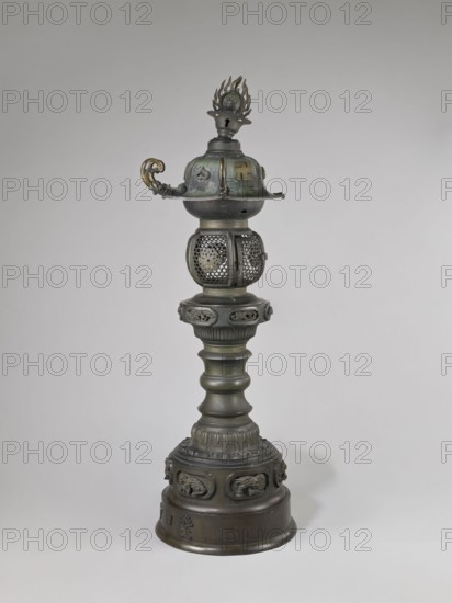 Temple lantern, Japanese, Edo period Man'en, 1860–1861; 1860, 9th month of the Year of the Monkey. Bronze; lantern on a high, multi-segmented shaft with a large round base; hexagonal roof decorated with dragons, pendants and bells at the eaves, topped with a flaming pearl. Assembled from elements cast in bronze, with relief and applied solid decorations. Iconography: dragons, swastika, monkey zodiac; function: temple lantern. Height 184.0 cm; weight 54.0 kg., A tall metal object stands against a plain light gray background, showing a vertical form made of a dark greenish-bronze metal with areas of darker patina and hints of muted gold. The top section features a flame-shaped finial above a rounded cap with a curved decorative handle on one side that has a more golden color. Below that is a spherical cage-like element with perforated decorative patterns. The central shaft is composed of multiple rounded and fluted segments with a ribbed collar, all in the same dark metallic green and br...