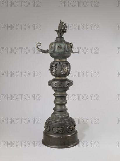 Temple lantern, Japanese, Edo period Man'en 1860–1861, 9th month of the Year of the Monkey, 1860. Cast bronze lantern assembled from elements with relief and applied solid decorations. Lantern on a high, multi-segmented shaft with a large round base; hexagonal roof decorated with dragons, pendants and bells on the edges, topped with a flaming pearl. Iconography: dragons, swastika, monkey zodiac. Function: temple lantern lamp. Weight 54.0 kg; height 184.0 cm., A tall metal ornamental object with a rounded base and a fluted column rising to a decorative midsection and an openwork spherical lantern, topped by a domed cap with curled handles and a flame-shaped finial; the object appears metallic with visible colors including dark gray, greenish patina, and hints of brass-like gold.