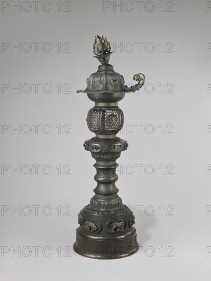 Temple lantern, Japanese, Edo period 1603–1868, Man'en 1860–1861. Bronze lantern featuring dragons, a swastika motif, and monkey zodiac imagery. Lantern on a high, multi-segmented shaft with a large round base. Hexagonal roof decorated with dragons, pendants and bells at the edges, topped with a flaming pearl. Assembled from elements cast in bronze with relief and applied solid decorations. Dated 1860 Man'en 1, 9th month, Year of the Monkey. Varia; lamp. Weight: 54.0 kg; height: 184.0 cm., A tall ornate metal object stands against a plain light gray background, showing a dark bronze to greenish patina with areas of muted gold; the piece has a rounded base with decorative raised panels and sculpted relief motifs, a flared tier above with vertical ridges, a segmented column with bulbous contours, a ringed section with carved details, a perforated spherical middle section with an openwork lattice, a wider bowl-like element topped by a domed cap with curved flanges, a curled ornamental han...