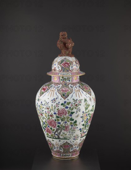 Guan vase with cover, ceramics, chrysanthemums, covers, Chinese art, Chinese culture, decorative vessel, lion iconography, peonies, Qing 1644–1911, ovoid belly, 19th century, China, vessel, height 92 cm, diameter 46 cm, A tall lidded ceramic vase with a glossy white base covered in floral and foliate painted decoration in pink, green, blue, yellow, and gold tones, featuring a group of pink peony-like flowers and green leaves arranged around the body, accented by smaller blue and yellow floral motifs and stylized scrolling patterns; the shoulder and foot have bands of pink and gold ornament with ornate cartouche-like panels in similar colors, and the lid is topped by a sculpted figure in a brownish-red glaze sitting on a domed section decorated to match the vase.