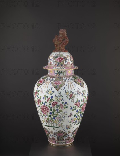 Guan vase with cover, ceramics; chrysanthemum and peony decoration with lion iconography; ovoid belly; Qing dynasty 1644–1911, 19th century; China; height 92 cm, diameter 46 cm;, A tall porcelain covered vase with a domed lid topped by a sculpted figure in a brownish-red color stands against a dark gray background; the body of the vase is predominantly white with an overall floral decoration featuring pink, green, blue, and yellow hues, with several large pink flowers and clusters of green leaves and smaller multicolored blossoms, accented by decorative cartouches and borders in pink and gold tones around the shoulder and base, and additional blue and green ornamental motifs dispersed across the surface.