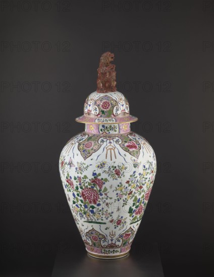 Guan vase with cover, ceramics, chrysanthemums, covers, Chinese art, Chinese culture, decorative vessel, lion iconography, peonies, Qing 1644–1911, ovoid belly, 19th century, China, vessel, height 92 cm, diameter 46 cm, The image shows a decorative lidded vase displayed against a dark gray background; the vase has a white base color with a floral pattern in pink, green, blue, yellow, and purple, featuring large pink blossoms and green leaves scattered among smaller multicolored flowers and foliage, and patterned ornamental bands in pink and gold near the neck and base; the lid echoes the floral motif and is topped by a sculpted reddish-brown figure; overall the object presents glossy ceramic surfaces with details in gold and touches of blue and green.