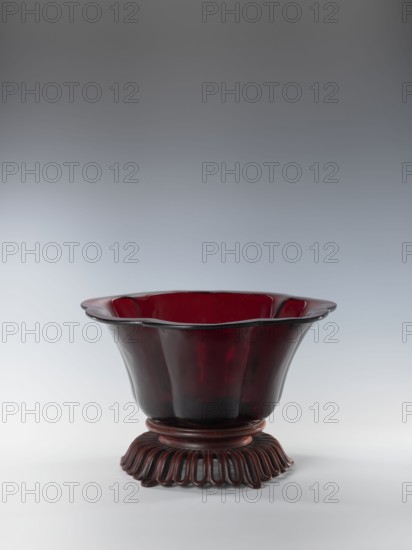 Bowl on base, bowls, Chinese art, Chinese culture, Qianlong style, Qing dynasty 1644–1911. Each segment slightly arched. The bowl's rim is rolled outward. Signed. Wooden base carved with an openwork pattern. 1736–1795, China. Vessel. Height 12.5 cm; width 26.8 cm., A single decorative bowl sits centered on a pale surface against a gradient gray background, the bowl flaring outward with a scalloped rim and smooth curved sides, made of translucent deep red material that darkens toward the base; it rests on a carved stand with a circular foot composed of repeated looped elements in a matching dark red-brown color.