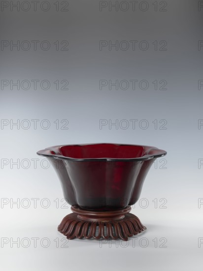 Bowl on base, bowl, Chinese art, Chinese culture, Qianlong style, Qing 1644–1911, Each segment slightly arched. The bowl's rim is rolled outward. Signed. Wooden base, carved with an openwork pattern. 1736–1795. China. Vessel. Height 12.5 cm; width 26.8 cm., A deep, translucent red glass bowl with a scalloped rim sits on a carved wooden stand, the bowl flaring outward with smooth, curved sides and a glossy surface that reflects light; the wooden stand is dark brown with an undulating, looped base and a rounded upper platform that supports the bowl, and the background is a smooth gradient of gray to light gray.