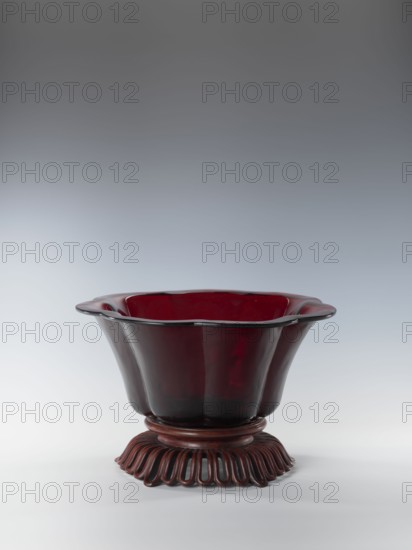 Bowl on base, bowl, Chinese art, Chinese culture, Qianlong style, Qing 1644–1911, Each segment slightly arched. The bowl's rim is rolled outward. Signed. Wooden base, carved with an openwork pattern. 1736–1795. China. Vessel. Height 12.5 cm; width 26.8 cm., A glossy red vessel with a fluted, wavy rim sits on a decorative base; the vessel's upper interior and exterior show deep red tones with darker accents and a subtle gloss, while the base is a darker reddish-brown with a carved, looped pattern. The background is a smooth gradient of gray transitioning from darker at the top to lighter toward the bottom, and the surface beneath the object is a pale gray-white.