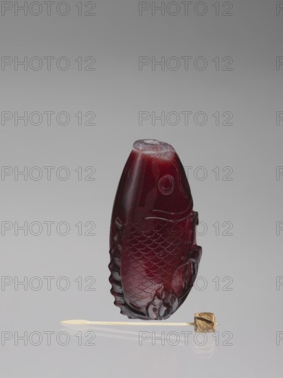 Snuffbox with spoon, shaped like a fish with a folded fin; decorated with bottles and carps; Chinese art; Chinese, Qing dynasty 1644–1911; 18th century; snuffbox vessel; height 7.0 cm, width 4.0 cm; provenance unknown., A carved fish-shaped object made of dark red translucent material with carved scale and fin details sits upright on a reflective surface, accompanied by a small pale stick with a light-colored pointed end and a small irregular tan-brown cube attached near the other end; the background is a smooth gradient of gray tones.