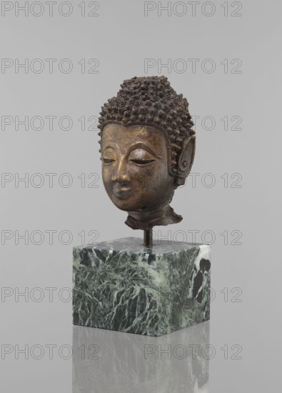 Buddha head - fragment of a sculpture, Buddhist art, Buddhism, art of Southeast Asia, Thai art, statue fragment with ushnisha, head, 12th–19th century, Thailand, figurine, height 11.5 cm, diameter 9.0 cm, A sculpted bronze head with a textured hair surface of small rounded curls and a serene facial expression sits mounted on a metal rod above a green and white marbled block; the head shows smooth, rounded facial features with closed eyes, arched eyebrows, a straight nose, and a small smiling mouth, and the ears are elongated with carved inner details; the bronze surface displays mottled tones of brown and dark gold with areas of darker patina, while the base is predominantly green with lighter green and white veining and a glossy finish, all set against a plain light gray background.