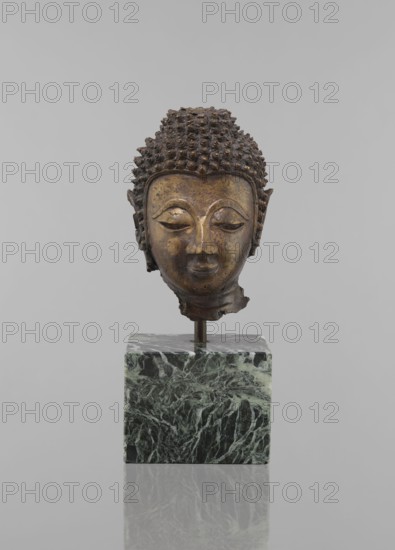 Buddha head - fragment of a sculpture, Buddhist art, Buddhism, art of Southeast Asia, Thai art, statues, fragments, ushnisha, heads, 12th–19th century, Thailand, figurine, height 11.5 cm, diameter 9.0 cm, A bronze-colored sculpted head with textured curly hair and a serene facial expression sits mounted on a metal rod above a green and black marbled cubic base; the background is a uniform light gray, and the surface beneath the base reflects the base faintly.