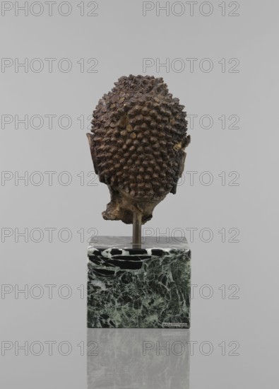 Buddha head — fragment of a sculpture, Buddhist art, Buddhism, art of Southeast Asia, Thai art, statue fragment, ushnisha, head, 12th–19th century, Thailand, figurine, height 11.5 cm, diameter 9.0 cm, A sculpted head viewed from the back mounted on a rectangular base, with a textured surface of rounded nodules across the head that are dark brown and bronze in color, and small exposed areas of lighter brown; the ears are visible at the sides with similar brown tones and some irregularities in the material; the mounting rod is metallic and dark, attached to a blocky base that has a marbled pattern in shades of green, black, and pale gray; the background is a uniform light gray and the surface beneath the base shows a soft reflection.