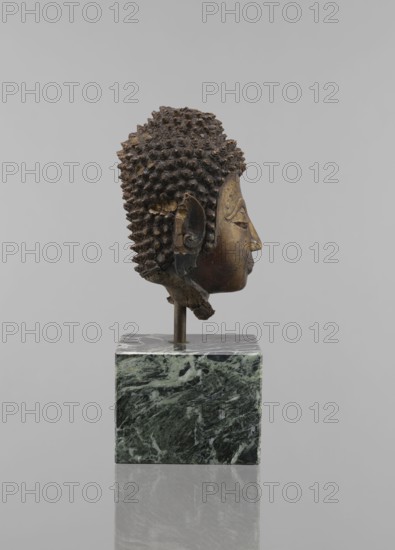 Buddha head — fragment of a sculpture; creator, Buddhist art, Buddhism, Southeast Asian/Thai art; statue fragment showing ushnisha and head; figurine; provenance, 12th–19th century; height 11.5 cm, diameter 9.0 cm., A sculpted head of a figure shown in profile mounted on a vertical rod and a square block base, with textured hair composed of small rounded curls and a smooth face with stylized features including a closed eye, a defined nose, and a small mouth; the sculpture shows areas of wear and roughness around the ear and lower neck. The visible colors include dark brown and bronze tones on the head, darker almost black areas in the hair, a green and white marbled pattern on the square base, and a neutral light gray background.