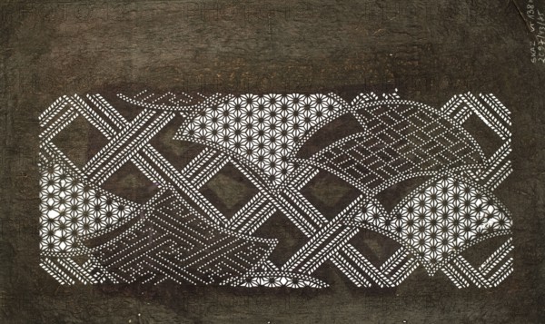 Katagami dyeing stencil with a modified wave pattern and diagonal check motif kawari seigaiha ni naname kôshi, Meiji period 1868–1912, katagami stencil, geometric motifs including seigaiha, asanoha, sayagata, nikuzushi; 19th/20th century Meiji period, Japan; stencil; height 40.0 cm, width 24.4 cm., The image shows a textured dark brown background with a central horizontal rectangular area made up of an intricate cutout pattern revealing a white layer beneath; the cutout forms repeating geometric motifs including star-like floral shapes and interlaced lattice bands, creating a layered mosaic of arc and diamond shapes. The visible colors are dark brown, black tones from the textured surface, and white from the underlying cutout areas.