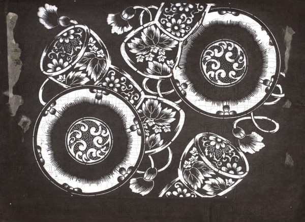 Dyeing stencil katagami with tsuzumi drum motif, Meiji period late 19th century, 1868–1912, Japanese; ise-katagami stencil, chuban format; motif: tsuzumi drum and floral ornaments; height 40.6 cm, width 30.5 cm., The image shows a high-contrast pattern of ornate dishware and floral elements arranged on a dark background, with a few large circular plates and several cups overlapping and interwoven with curved handles and stylized flowers. The dishware and floral designs are rendered in stark white against deep black, creating bold outlines and detailed decorative motifs with petal and leaf shapes, scalloped rims, and textured center medallions. The background is textured black with irregular worn or torn patches along the edges that appear in lighter gray tones, and there are fine crackle-like lines within the white areas of the pattern. The visible colors are black, white, and shades of gray.