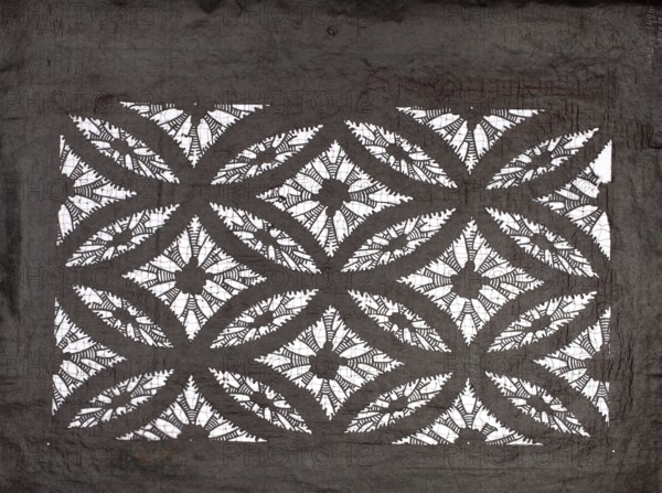 Katagami dyeing stencil with a motif of overlapping circles shippô. Ise-katagami stencil, chuban format. Pattern: geometric motif—rhombuses inscribed in ellipses with stylized floral rosettes in the rhombuses. Late 19th century Meiji period, Japan. Stencil katagami. Height 41.0 cm, width 31.5 cm., A rectangular patterned fabric with a dark gray background and white cutout areas forming a repeating geometric floral motif, featuring a grid of diamond shapes each with a central flower-like element surrounded by petal-shaped cutouts and curved leaf-like arcs; the dark gray fabric shows visible creases and texture around the patterned area, and the white cutouts reveal a contrasting bright surface beneath.