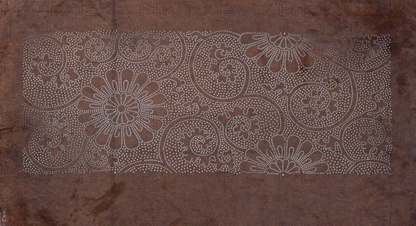 Dyeing stencil katagami with a plant tendril and chrysanthemum motif karakusa ni kiku, ise-katagami stencil, chuban format; flowers, chrysanthemums, plant tendrils karakusa pattern; late 19th century Meiji period, Japan; height 41.8 cm, width 25.8 cm., The image shows a rectangular section of a textured surface with a pattern of many small circular perforations forming floral and swirling designs; the background surface is various shades of brown with darker and lighter brown mottling, and the perforations reveal a lighter color that appears off-white or cream.