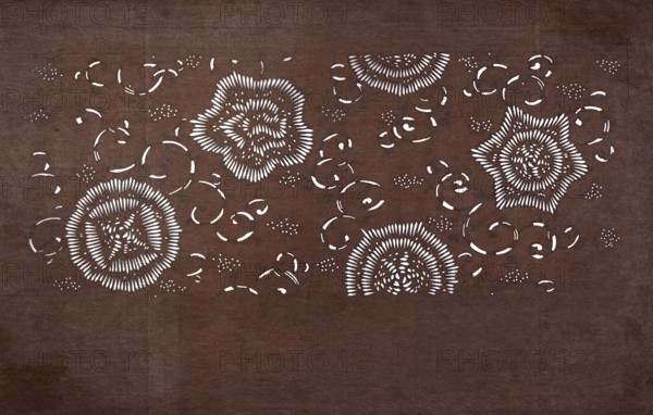 Katagami dyeing stencil with a motif of stylized flower rosettes and plant tendrils kawari kamon ni karakusa moyô, Japanese, Meiji period 1868–1912, ise-katagami stencil, chuban format, floral ornaments, karakusa pattern, 19th/20th century Meiji period, Japan; stencil; height 41 cm, width 25.1 cm., A textured brown background fills the image with a pattern of cutout shapes revealing white beneath; the cutouts form a few concentric circular and floral motifs with radiating dash-like elements, several star-like floral outlines, and a scattering of curved arc shapes and small dot clusters; the visible colors are brown and white.