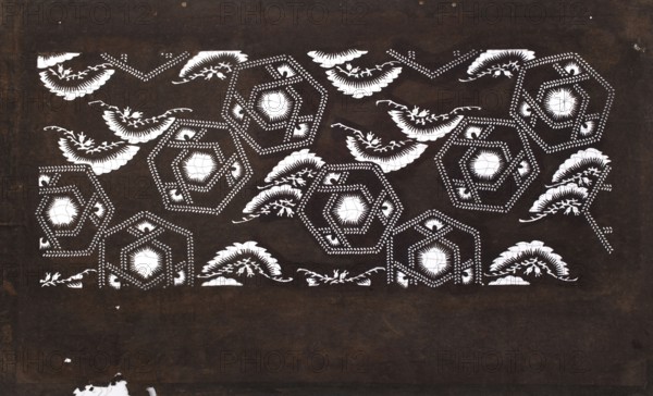 Dyeing stencil katagami with a pine tree and stylized hexagon motif matsu ni kawari kikkô moyô; unknown creator; Meiji period 1868–1912, 19th/20th century Japan; ise-katagami stencil, chuban format; geometric and plant patterns, pine trees; height 40.5 cm, width 24.5 cm., The image shows a patterned rectangular panel with a dark brown background and a repeating white decorative motif. The motif includes a group of hexagonal shapes outlined with dotted and chevron-like white markings, each with a central circular white area, and a few stylized white leaf- or feather-like forms interspersed among the hexagons. The white shapes have some fine jagged and scalloped edges and small branch-like extensions. The dark brown background appears textured with subtle variations in shade and a few small worn or chipped areas where the dark brown gives way to lighter tones.