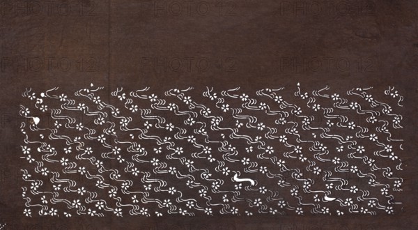 Katagami dyeing stencil with cherry blossom and flowing water motif sakura ni ryûsui, katagami stencil, chuban format; stencil of water, plant patterns, flowers, cherries sakura; 19th century Edo period, Japan; height 40.8 cm, width 22.3 cm., A dark brown textured background fills the image with a horizontal band near the bottom composed of a repeating white floral and wavy motif; the band contains a group of small stylized flowers and flowing curved lines in white that form a dense patterned area against the brown, with occasional isolated white shapes punctuating the pattern.