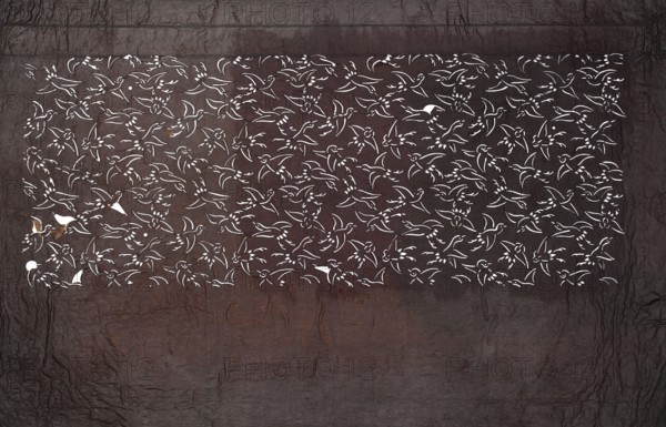 Katagami dyeing stencil; pattern of densely arranged birds in flight, ise-katagami stencil, Meiji period 1868–1912, late 19th–early 20th century, Japan; stencil; height 39.7 cm, width 24.7 cm; Japanese art, stencils., A rectangular section filled with a repeating bird motif cut out in white sits against a textured dark brown and gray background; the birds are stylized with simple outlines and wing shapes, appearing in a continuous pattern across the rectangle, and there are a few small areas where the white cutouts reveal underlying rust-colored patches; the surrounding surface shows crinkled, leathery texture in shades of deep brown, charcoal gray, and muted reddish-brown.