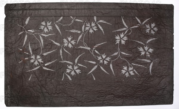 Katagami dyeing stencil; wild carnation pattern; maker, Japanese; Meiji period 1868–1912; ise-katagami stencil; floral ornament; 19th/20th century; stencil; height 40.6 cm, width 25.1 cm., A textured dark brown rectangular sheet with a crinkled surface fills the image, featuring a pattern of floral and leaf shapes created by narrow horizontal cutouts that reveal a bright white background beneath; the cutout shapes form several stylized flowers with radiating petal-like elements and long slender leaves connected by curved stems, arranged across the sheet in a repeating, flowing layout; subtle variations in the brown surface show areas of slightly lighter and darker brown, and the outer edge of the sheet is irregular against the white border.