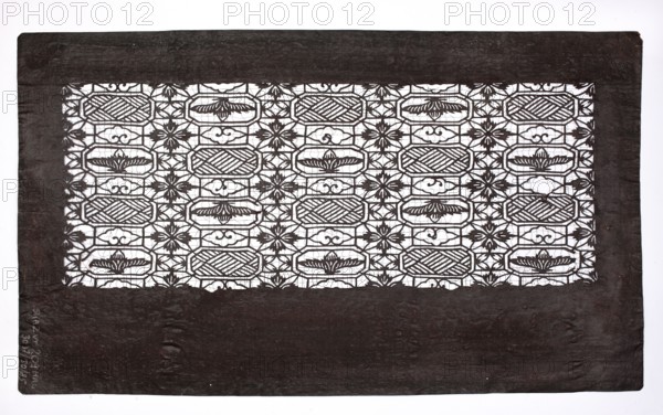 Katagami dyeing stencil; geometric-floral pattern stylized after the traditional Chinese brocade pattern syokko; maker, Japanese; Meiji period 1868–1912; ise-katagami stencil with brocade ornaments; 19th–20th century; stencil; height 40.3 cm, width 23.6 cm., A rectangular sheet with a dark border surrounds an inner panel of intricate cutwork forming a repeating geometric and floral pattern; the border is a deep brownish-black and shows subtle texture and creases, while the inner cutwork is dark against a bright white background, creating high contrast. The cutwork panel contains a grid-like arrangement of oval and octagonal shapes alternating with stylized floral motifs and small decorative curlicues, all rendered in the same dark color as the border, with areas of white visible through the openwork. The overall composition is flat and centered within a clean white surrounding, and the visible colors are dark brownish-black and white.