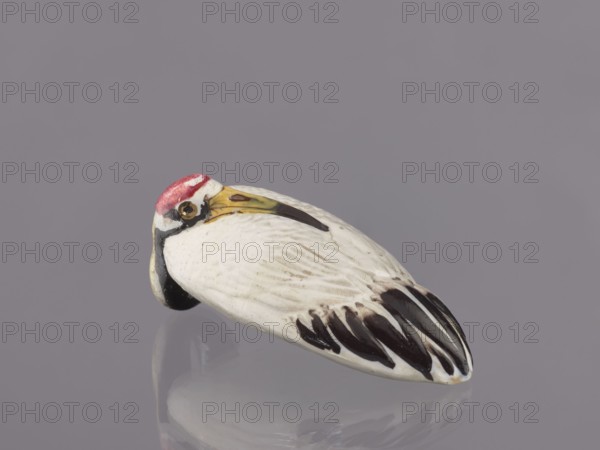 Vase, unknown ceramicist; Meiji period 1868–1912, Japan; ceramics; vessel vase; subject: cranes iconography; dimensions: height 9 cm, diameter 3.5 cm; 19th/20th century., A small ceramic bird figure resting on a reflective surface, showing a rounded body with carved feather details; its main body color is white with dark brown and black markings on the tail feathers and wing tips, a yellowish beak with a darker tip and a small dark eye area, and a red patch on the top of its head, all set against a muted grayish-lavender background.